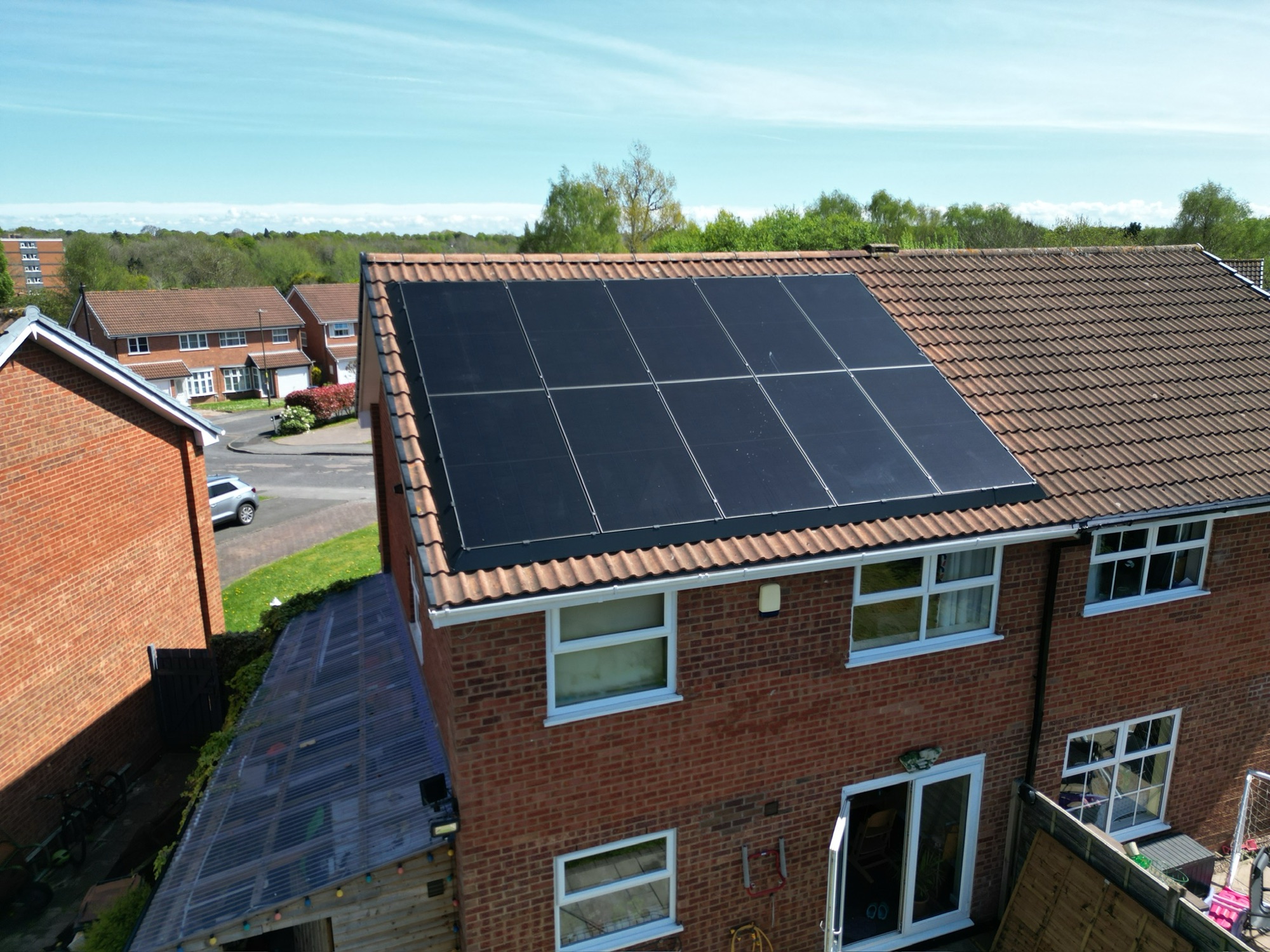 A twelve-panel all-black array on the tiled roof of a semi-detached house, photographed from above on a clear day.