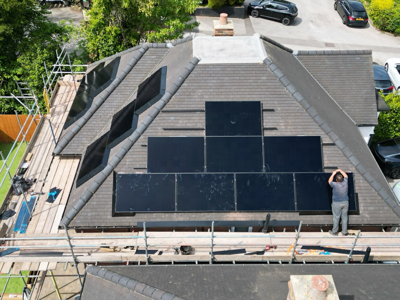An installer working across a hipped slate roof, panels part-laid and scaffold boards along the eaves.