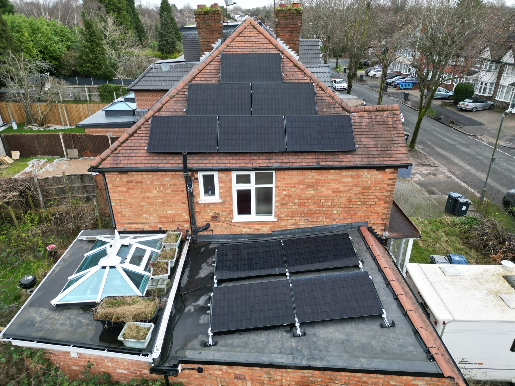 A red-brick house with panels on the main pitched roof and a second array on the flat roof extension below.